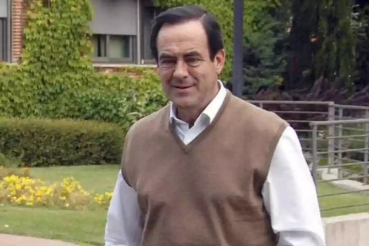 Hombre de pie al aire libre con un suéter marrón y camisa blanca, con un fondo de vegetación y un edificio.