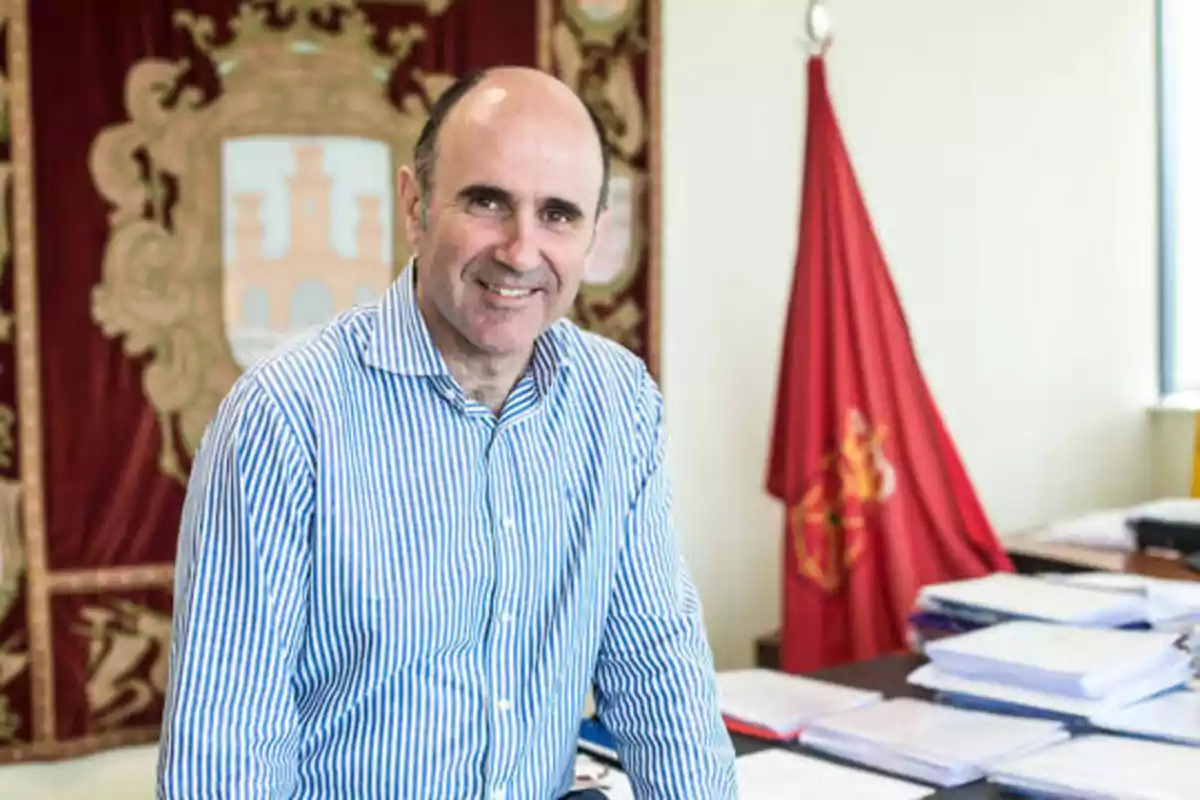 Hombre con camisa de rayas azules y blancas sonriendo en una oficina con una bandera roja y un escudo en el fondo.
