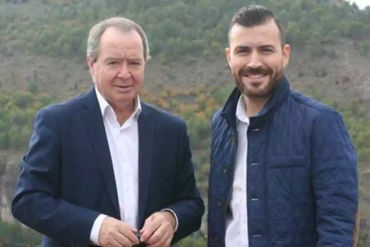 Dos hombres posando al aire libre con un paisaje montañoso de fondo.