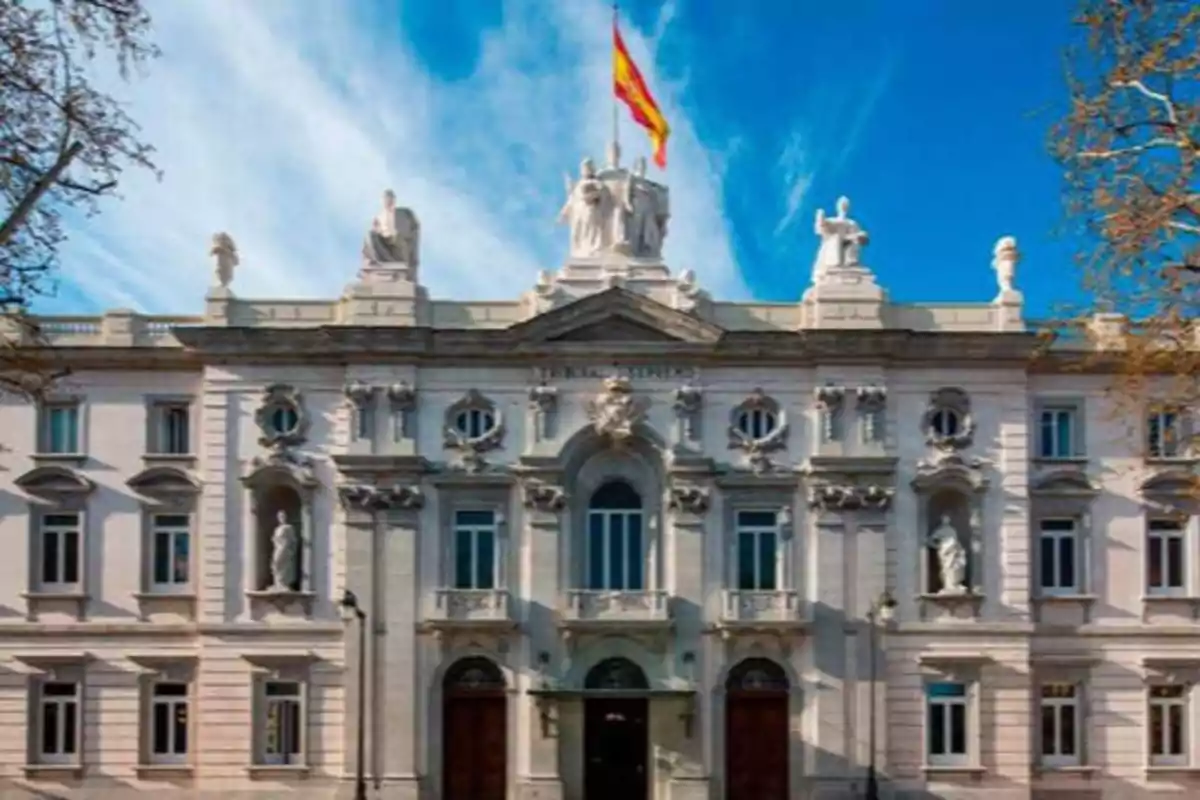 Fachada de un edificio histórico con una bandera ondeando en la parte superior y estatuas decorativas en el techo.