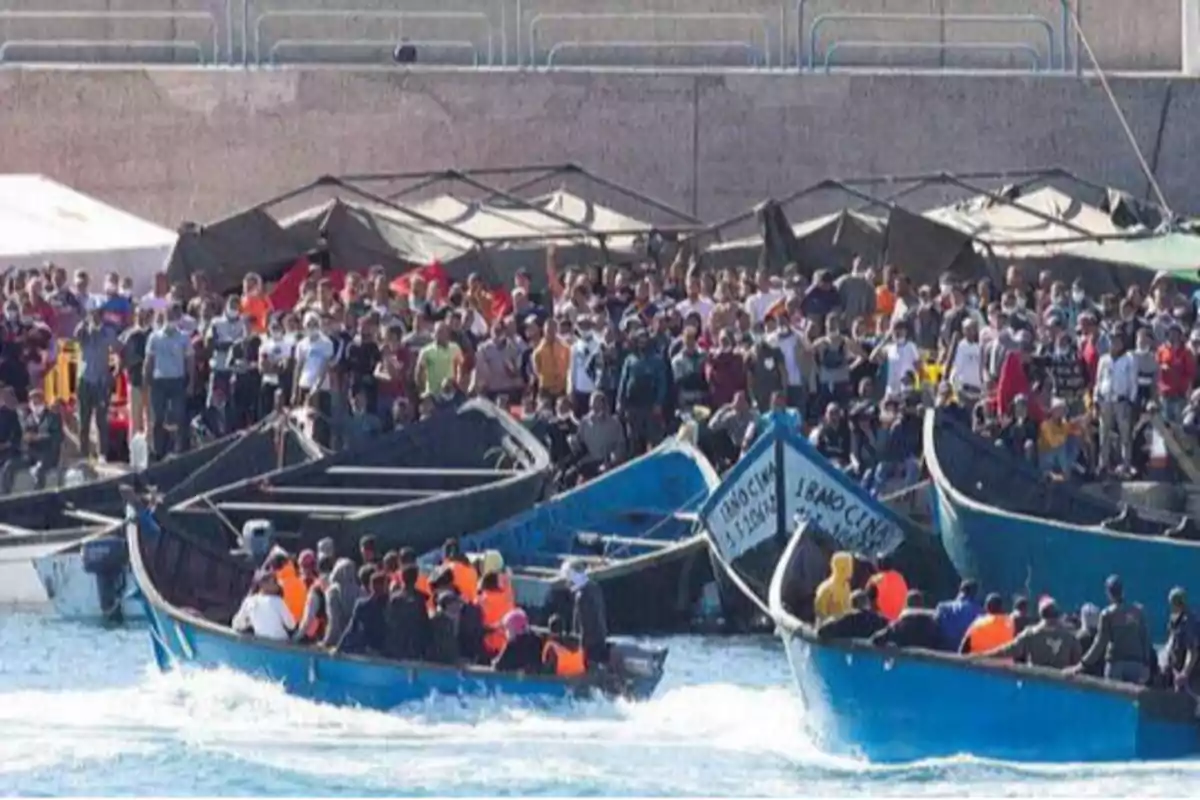 Una multitud de personas se encuentra en un puerto, con varias embarcaciones llenas de gente en el agua.