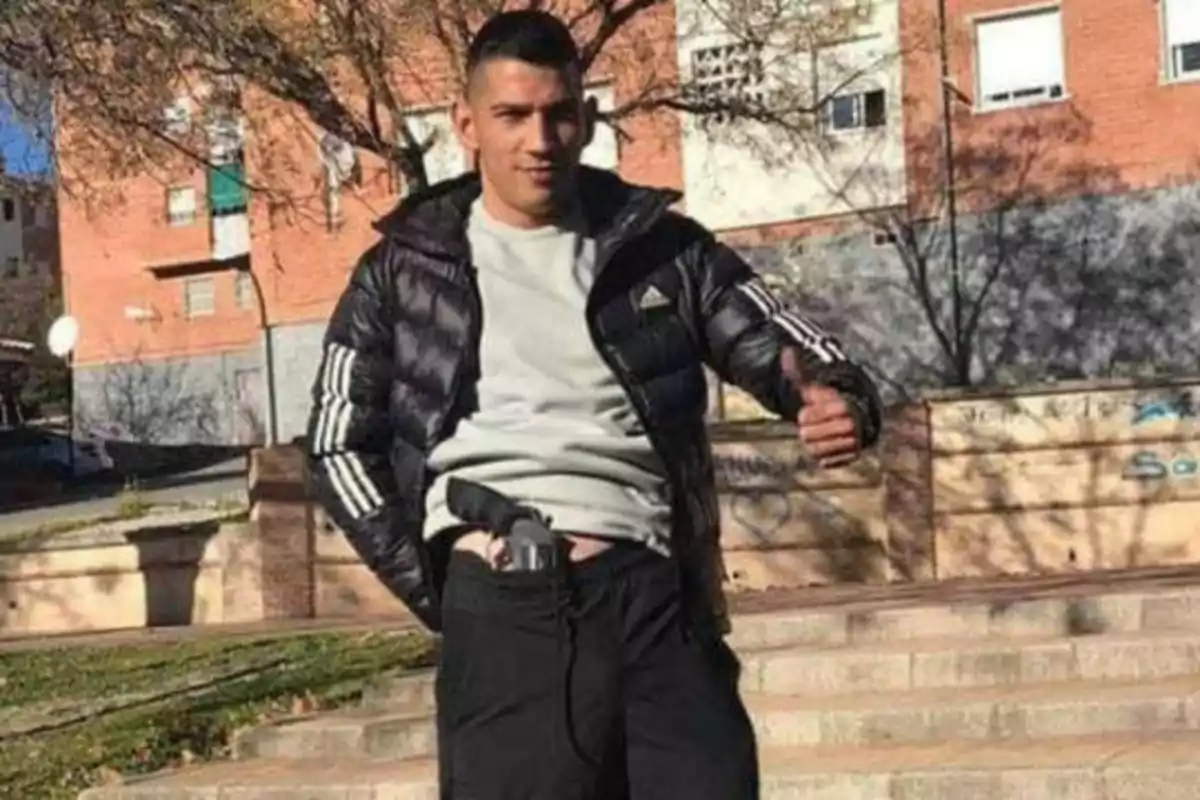 Un hombre con una chaqueta negra y una camiseta gris muestra un gesto de aprobación con el pulgar mientras sostiene una pistola en la cintura, con edificios de ladrillo y árboles al fondo.