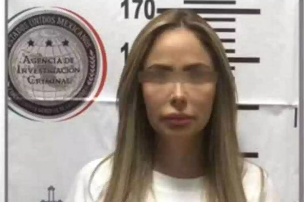 Una mujer con el cabello largo y lacio está de pie frente a una pared con una escala de altura y un emblema de la Agencia de Investigación Criminal de México.