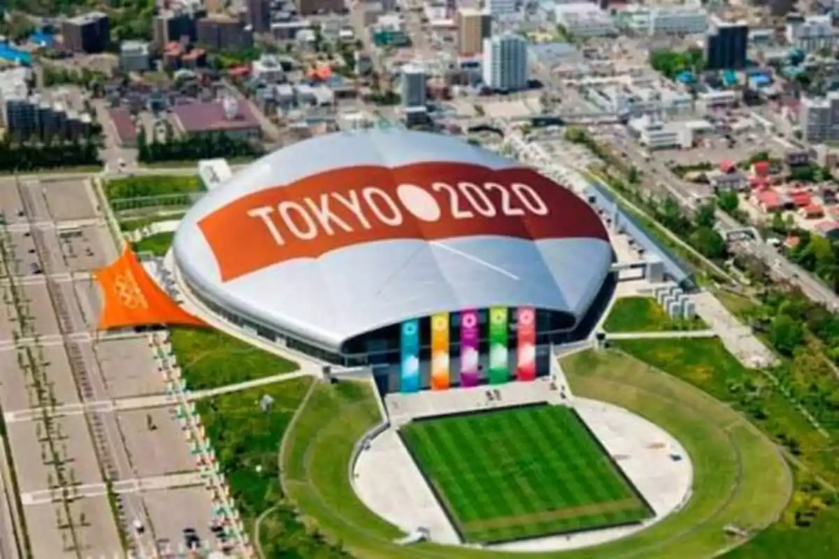 Vista aérea de un estadio con el logotipo de "Tokyo 2020" en el techo, rodeado de áreas verdes y edificios.