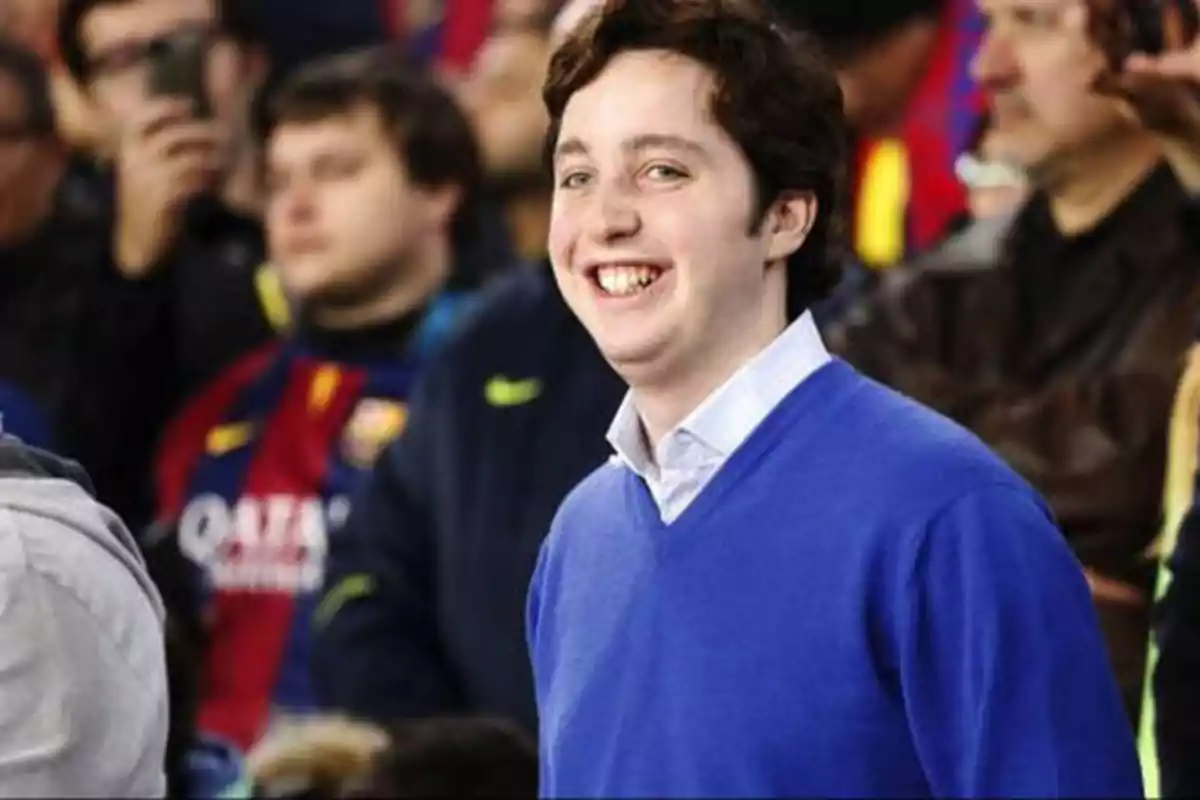 Un hombre sonriente con un suéter azul en un evento deportivo con personas de fondo.