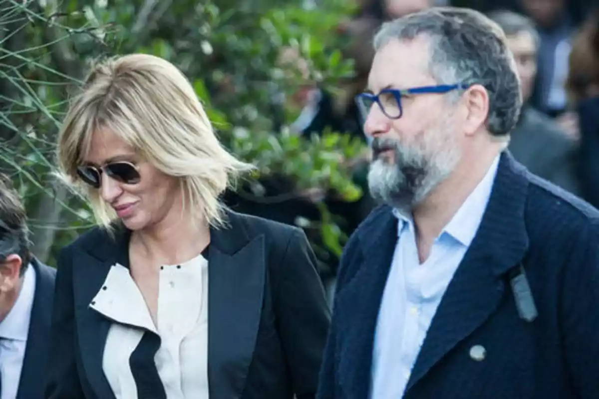 Dos personas caminando al aire libre, una mujer con gafas de sol y un hombre con barba y gafas.