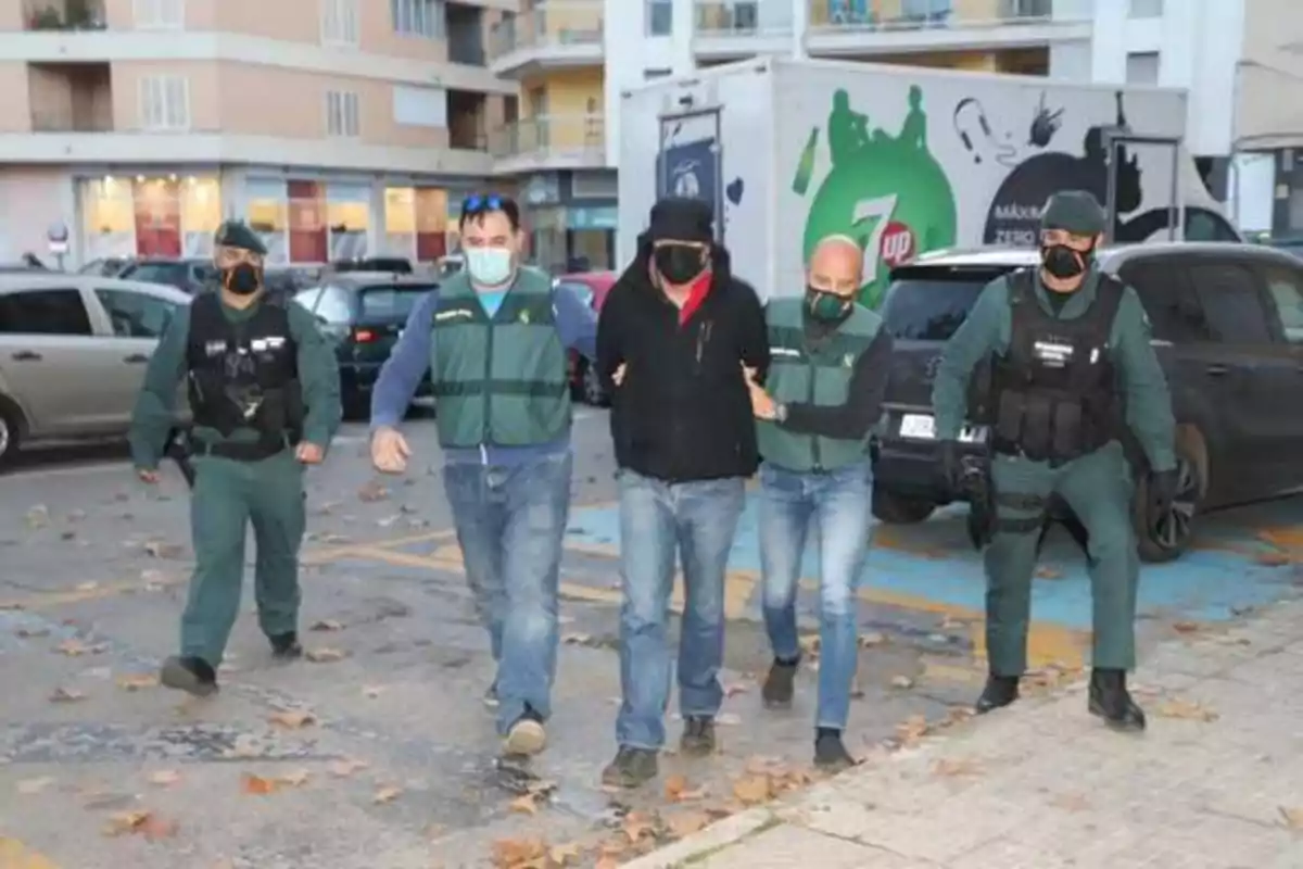 Un grupo de agentes de la Guardia Civil escolta a un hombre esposado en una zona urbana con varios vehículos estacionados y un camión de fondo.