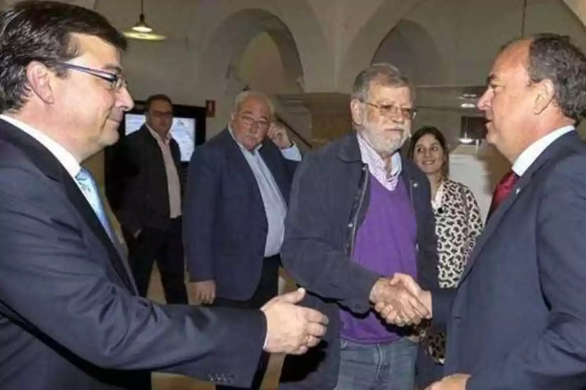 Varias personas en un evento formal, dos de ellas estrechándose la mano mientras otras observan en el fondo.