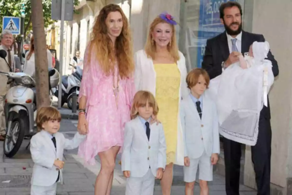 Una familia posando en la calle, con tres niños pequeños vestidos con trajes claros y corbatas, una mujer con un vestido rosa, otra mujer con un vestido amarillo y un hombre con traje oscuro sosteniendo a un bebé.
