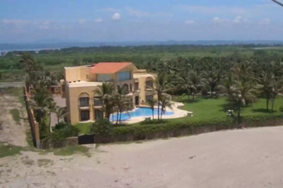 Una casa grande con piscina rodeada de palmeras y vegetación, ubicada cerca de una playa.