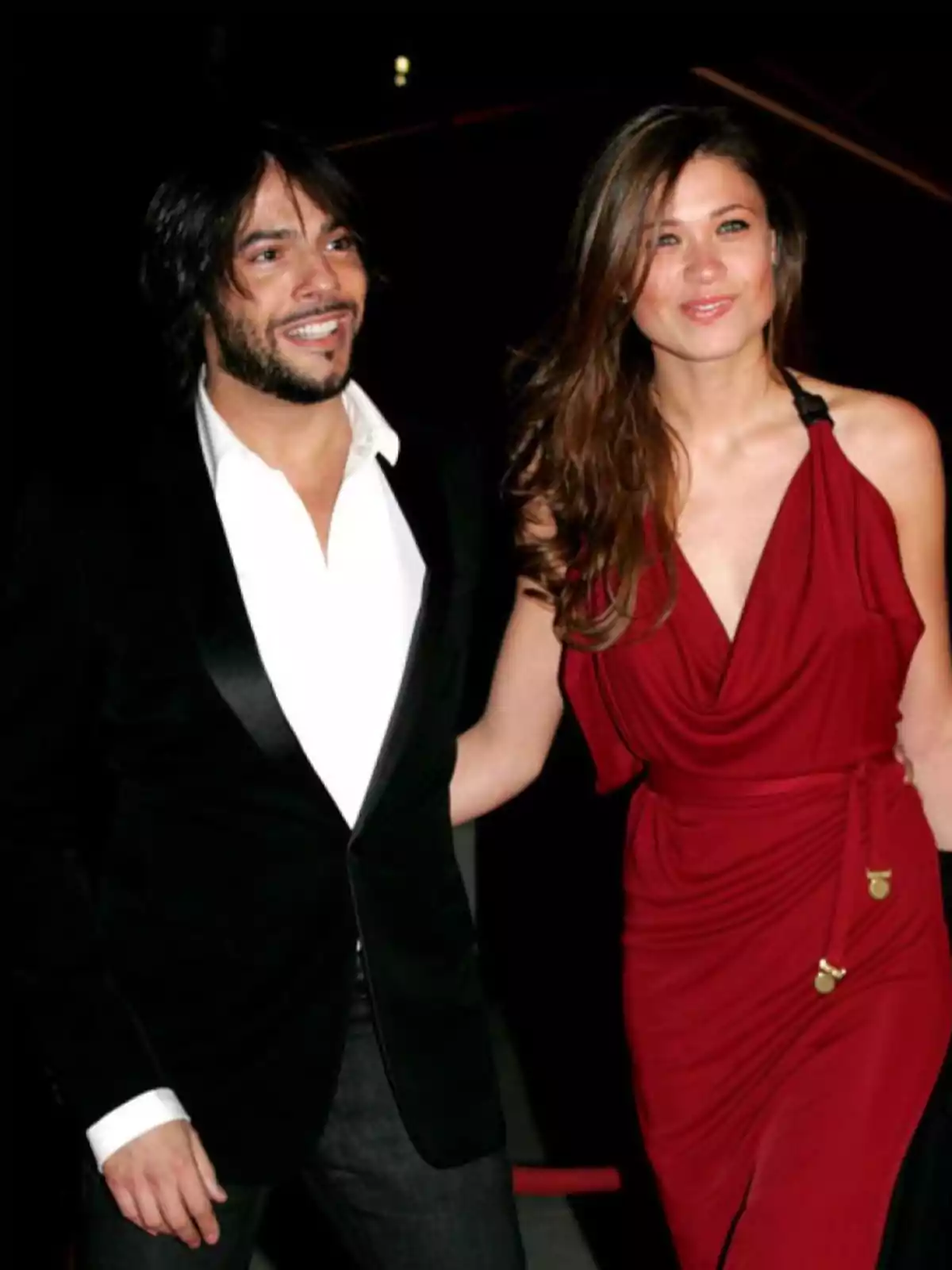Una pareja elegantemente vestida, el hombre con un traje negro y camisa blanca, y la mujer con un vestido rojo, posando juntos y sonriendo.