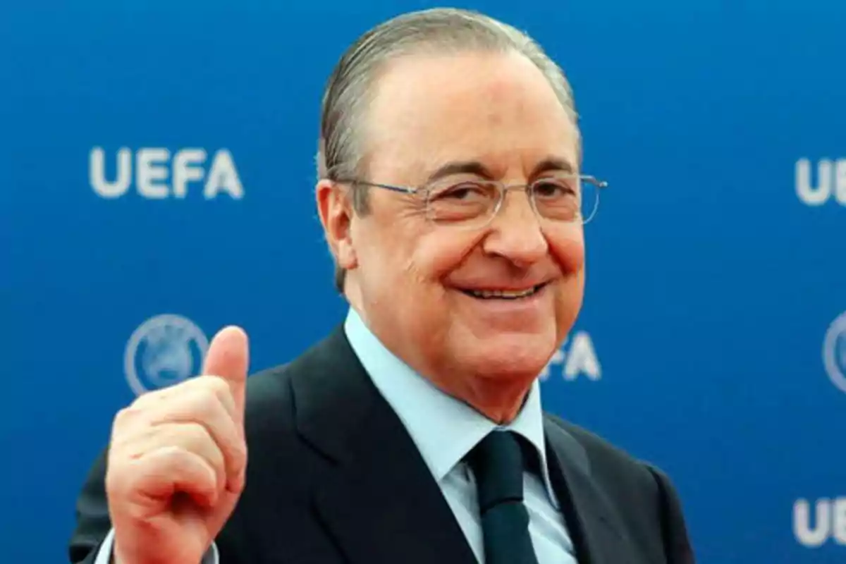 Hombre con gafas y traje oscuro sonriendo y levantando el pulgar frente a un fondo azul con el logo de la UEFA.