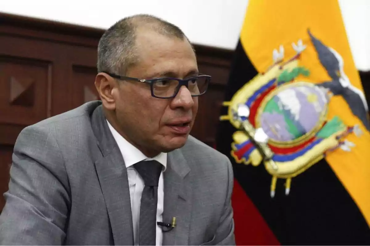 Hombre con gafas y traje gris sentado frente a una bandera de Ecuador.