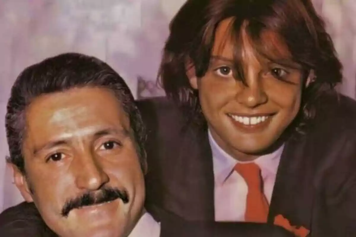 Dos hombres sonrientes posando juntos, uno con bigote y el otro con cabello largo y una corbata roja.