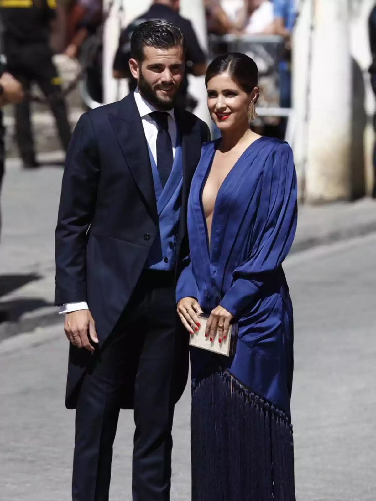 Una pareja elegantemente vestida, él con un traje oscuro y ella con un vestido azul profundo, posando juntos en un evento al aire libre.
