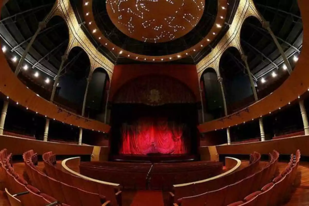 Un teatro con un escenario cubierto por una cortina roja, rodeado de asientos y con un techo decorado con luces.