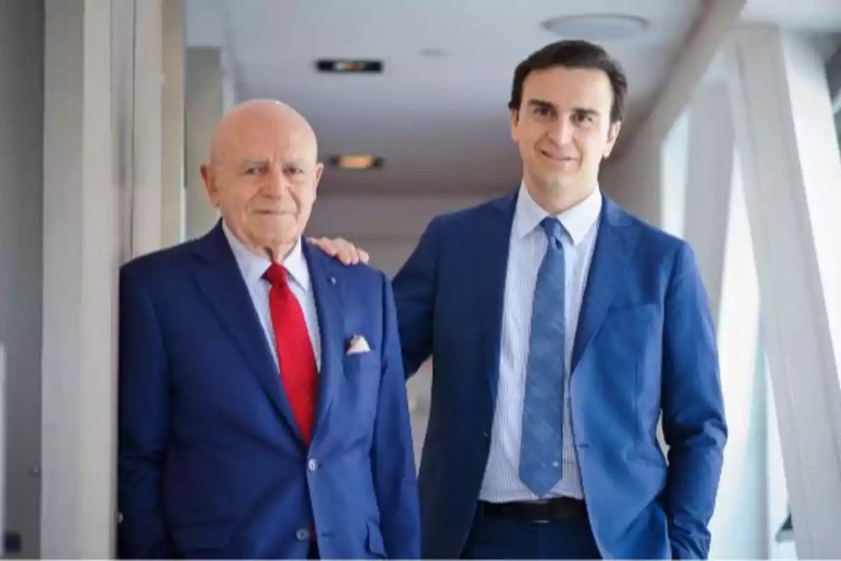 Dos hombres de traje azul posando juntos en un pasillo iluminado.