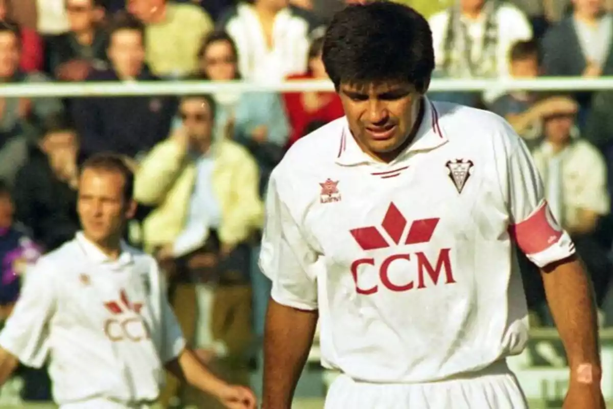 Un jugador de fútbol con uniforme blanco y brazalete de capitán en un campo de juego con espectadores al fondo.