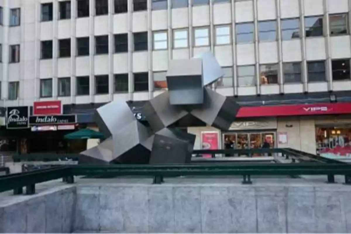 Escultura de cubos metálicos entrelazados frente a un edificio con ventanas y tiendas en la planta baja.