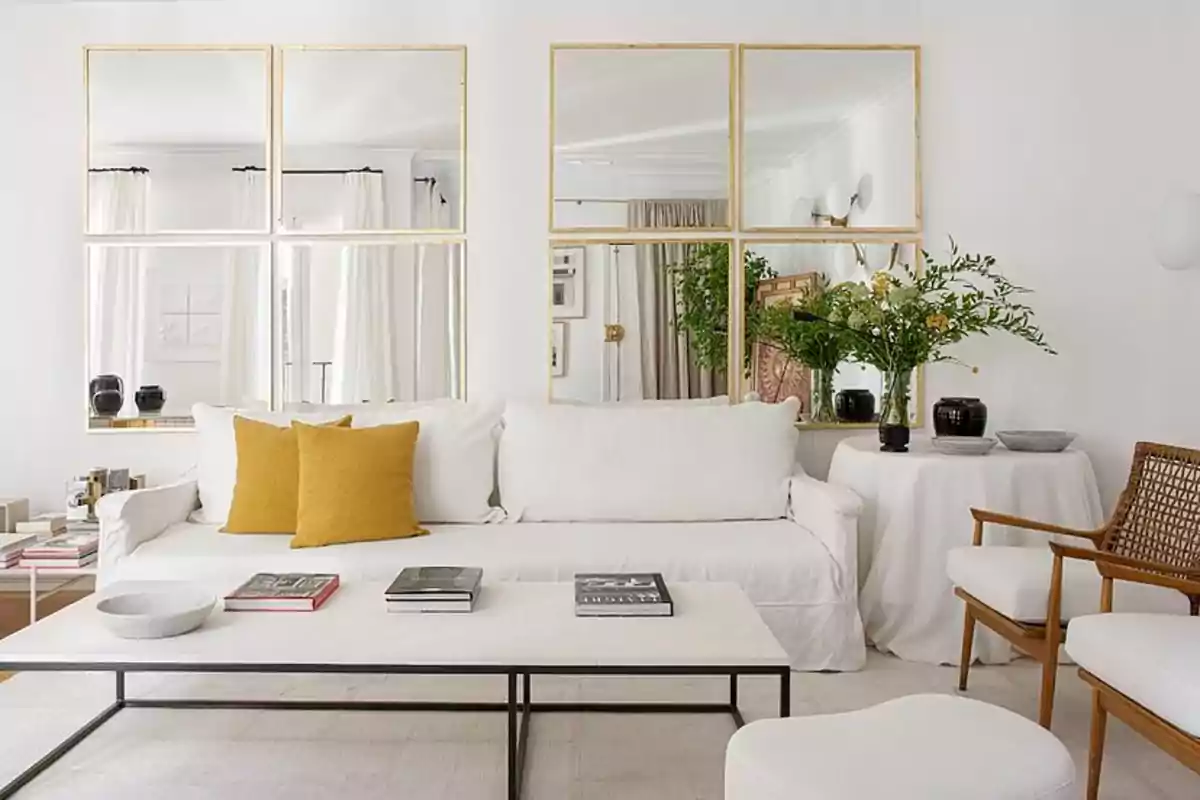Una sala de estar moderna y luminosa con paredes blancas, un sofá blanco con cojines amarillos, una mesa de centro blanca con libros y un cuenco, un espejo grande dividido en cuatro secciones en la pared, una mesa redonda con un mantel blanco y un jarrón con flores, y una silla de madera con cojines blancos.