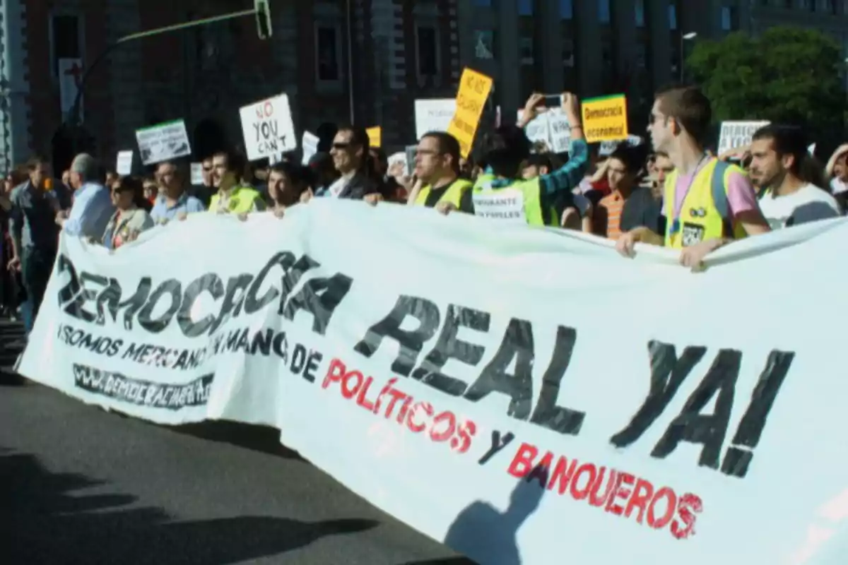 Un grupo de personas participa en una manifestación sosteniendo una gran pancarta que dice Un grupo de personas participa en una manifestación sosteniendo una gran pancarta que dice