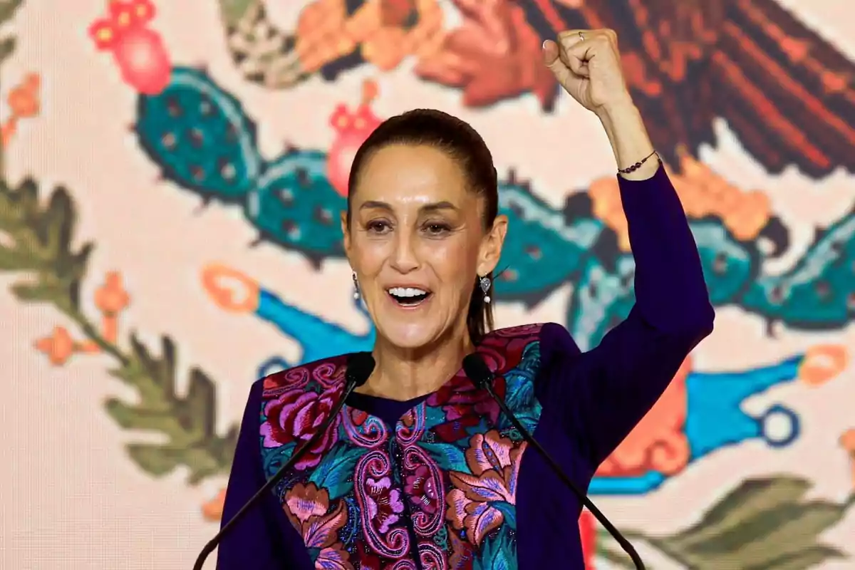 Una mujer con el puño levantado, vistiendo una blusa con bordados coloridos, habla frente a un micrófono con un fondo que parece ser el escudo nacional de México.