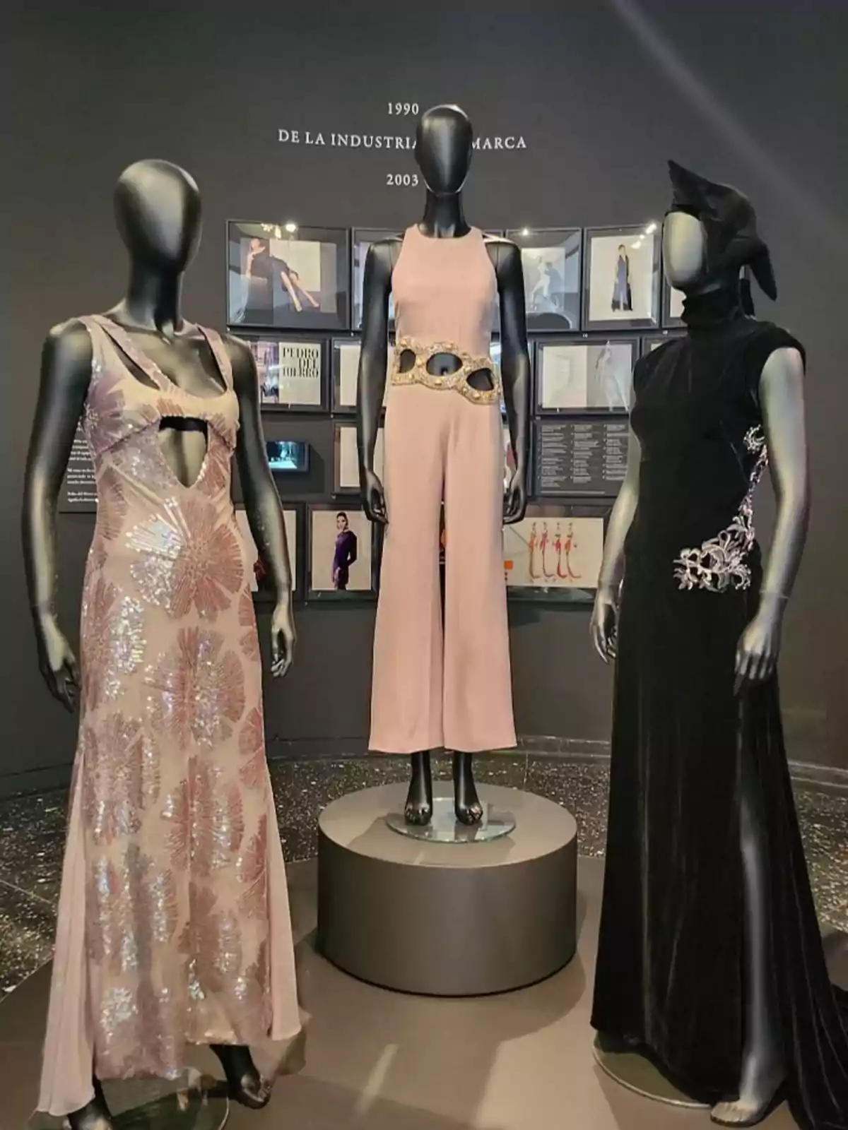 Tres maniquíes exhiben vestidos de alta costura en un museo de moda, con un fondo de fotografías y textos explicativos.