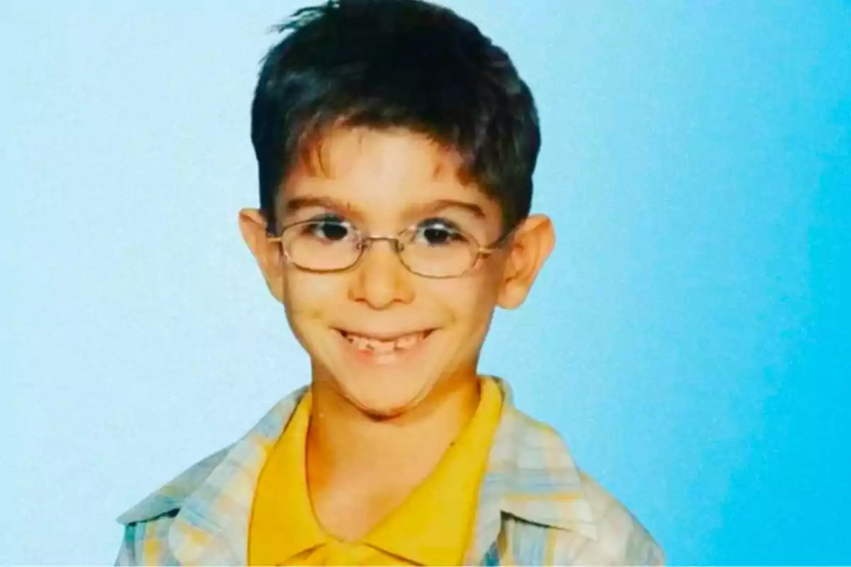 Un niño con gafas y una camisa amarilla sonríe frente a un fondo azul.