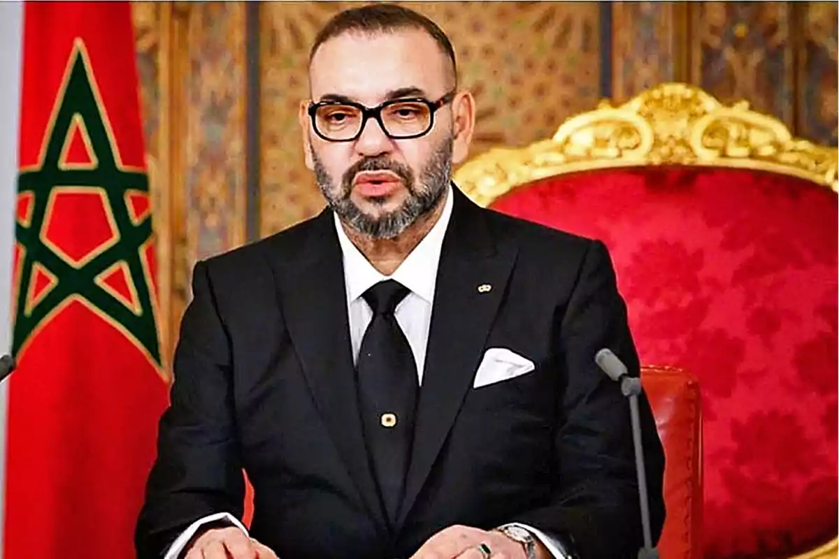 Hombre con gafas y barba, vestido de traje negro, sentado en un trono rojo con un fondo decorativo y una bandera con una estrella verde.