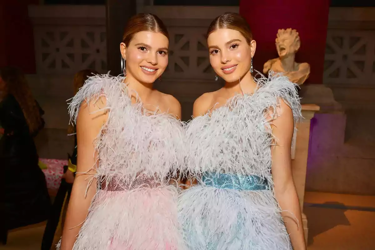 Dos mujeres con vestidos de plumas posando juntas y sonriendo en un evento elegante.
