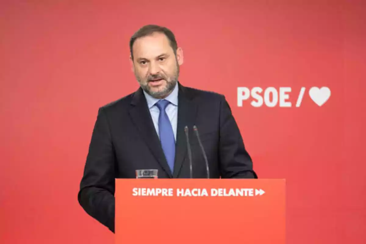 Hombre de traje hablando en un podio rojo con el logo del PSOE y el texto "SIEMPRE HACIA DELANTE" en el fondo.