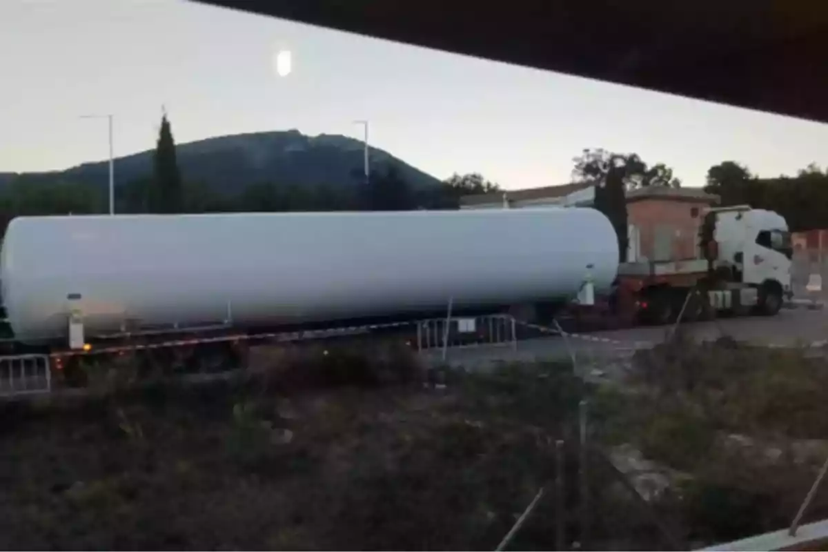 Un tráiler transporta un enorme tanque blanco por una carretera cerca de una zona rural con montañas al fondo. Un tráiler transporta un enorme tanque blanco por una carretera cerca de una zona rural con montañas al fondo.