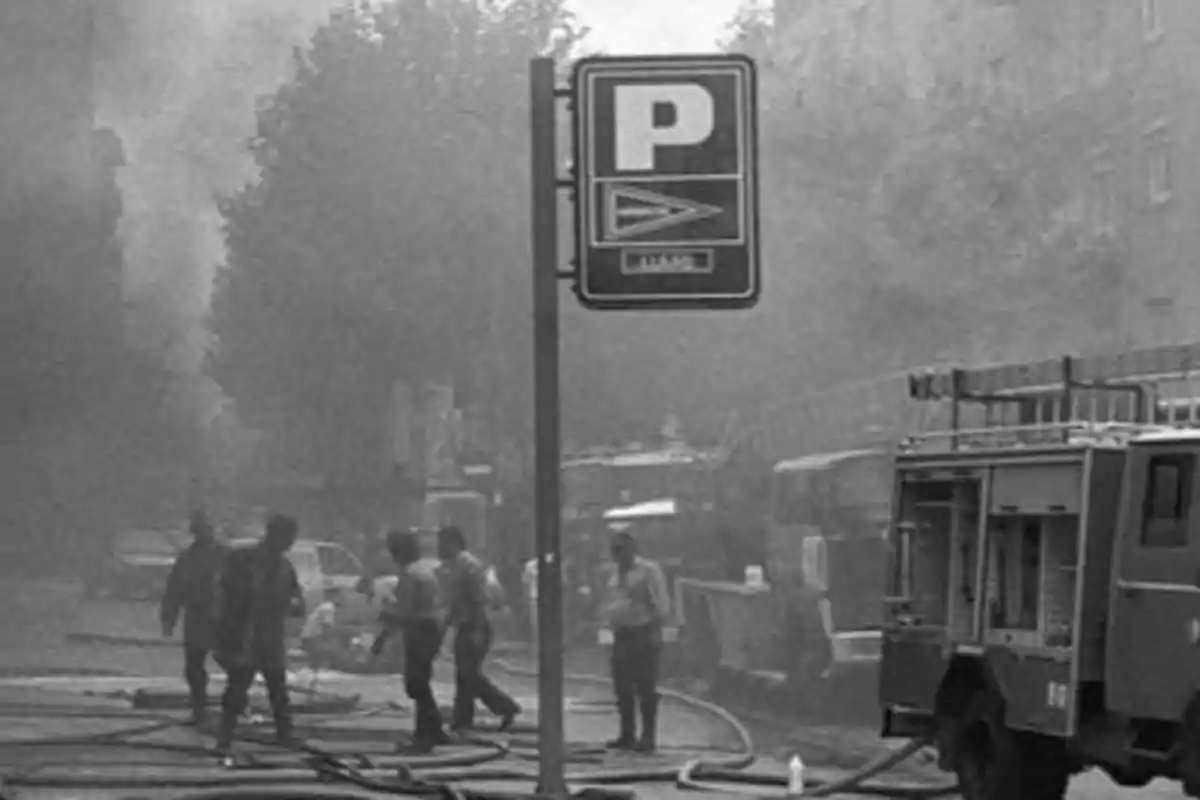 Bomberos trabajando en una calle llena de humo con una señal de estacionamiento visible.