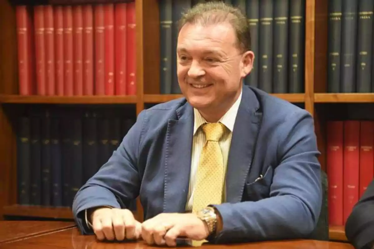 Hombre sonriendo sentado en una mesa con estanterías llenas de libros detrás.