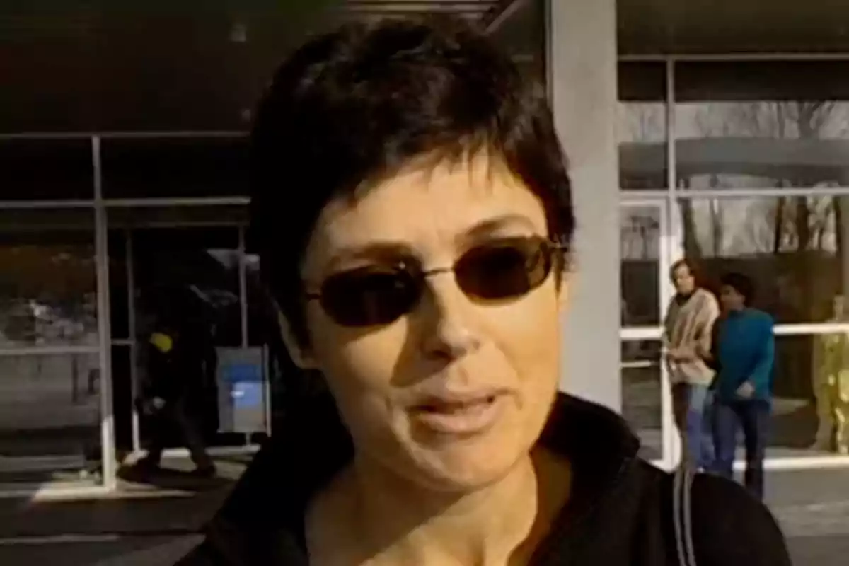 Mujer de cabello corto y oscuro usando gafas de sol frente a un edificio con personas al fondo Mujer de cabello corto y oscuro usando gafas de sol frente a un edificio con personas al fondo