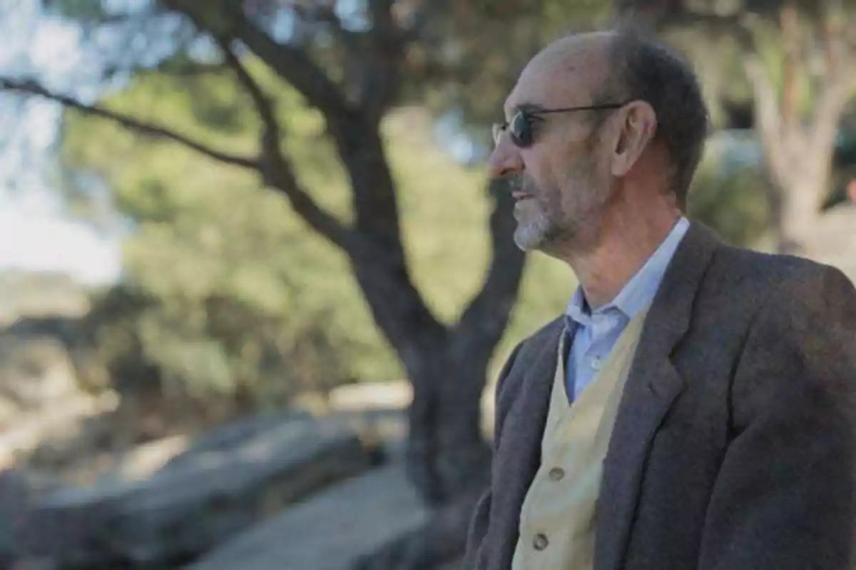 Un hombre mayor con gafas de sol y barba, vestido con un chaleco amarillo y una chaqueta marrón, está de pie al aire libre con árboles y un fondo natural desenfocado.