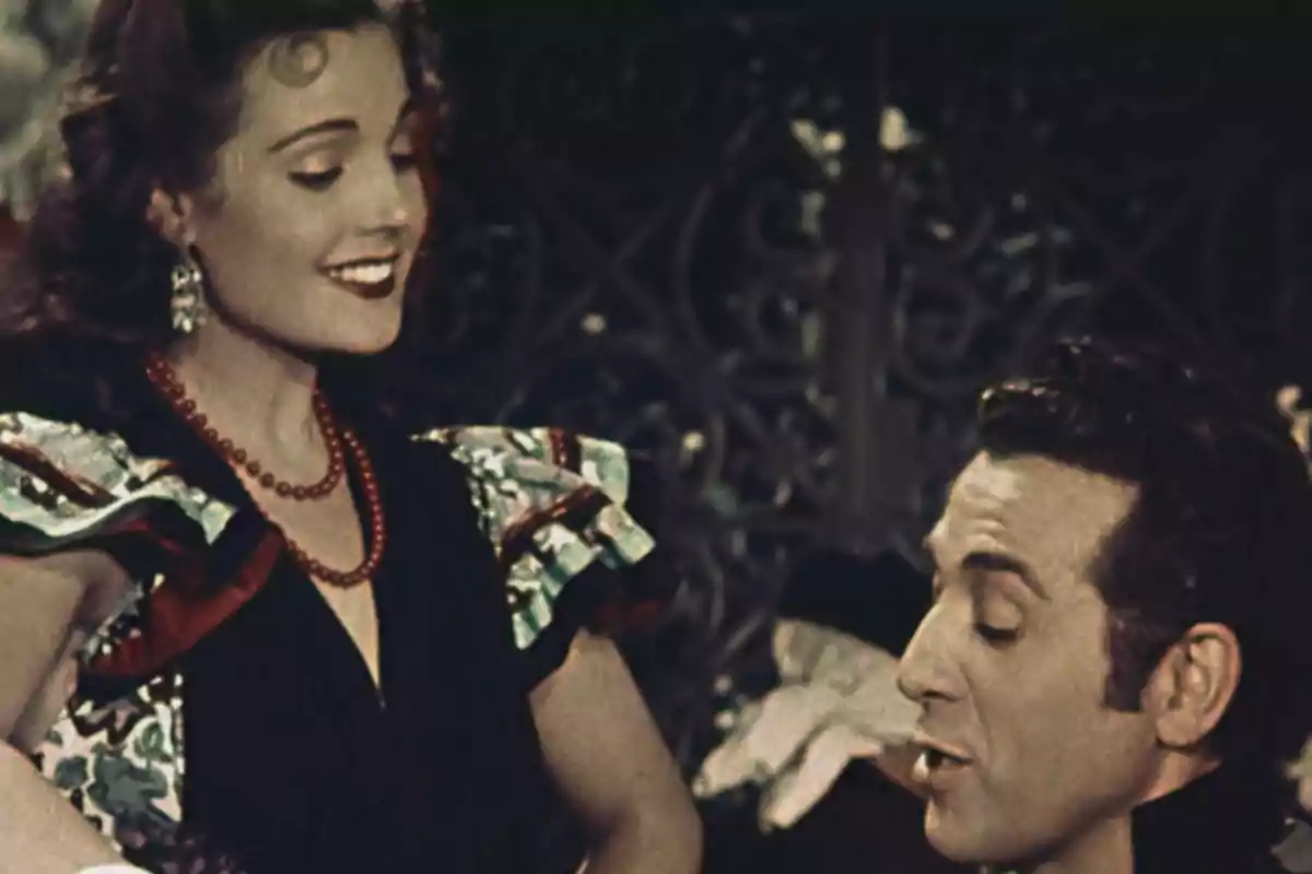 Una mujer sonriente con un collar de perlas rojas y un hombre hablando, ambos en un entorno decorado.