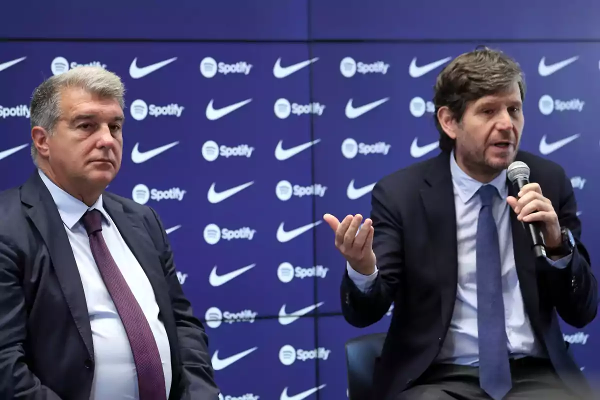 Dos hombres en trajes formales en una conferencia de prensa con logotipos de Nike y Spotify en el fondo.