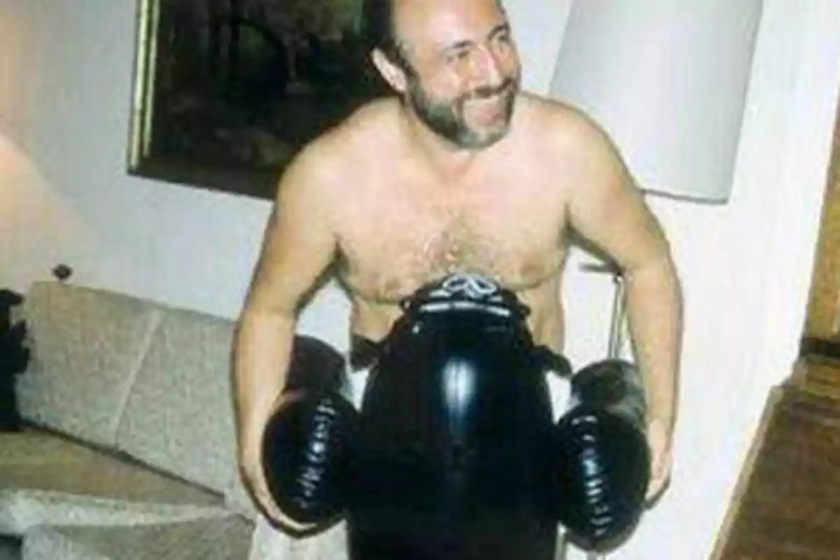 Hombre sin camisa sonriendo mientras sostiene un muñeco inflable de boxeo en una sala de estar.