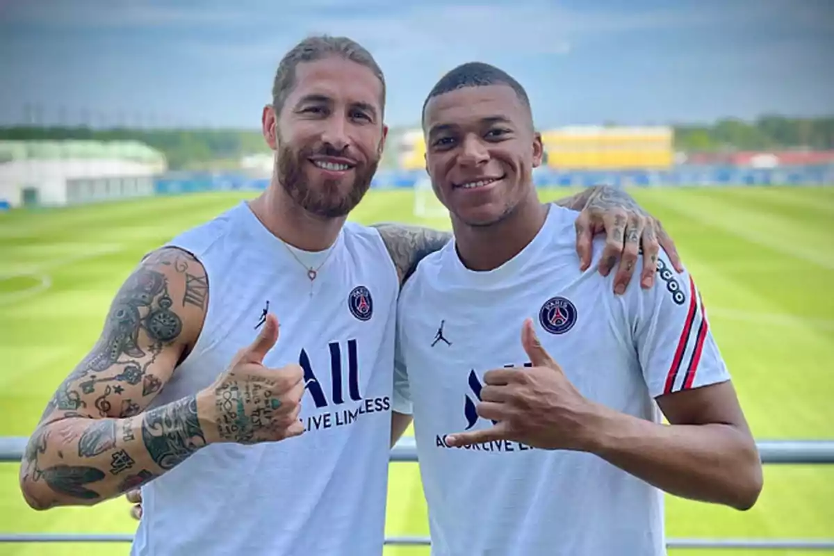 Dos jugadores de fútbol del PSG posando juntos en un campo de entrenamiento, ambos con el pulgar hacia arriba y sonriendo.
