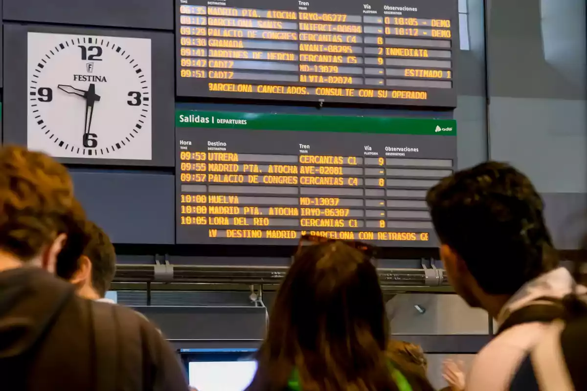 Personas observando un tablero de salidas de trenes en una estación, con un reloj grande mostrando la hora y varias pantallas con información de horarios y destinos.