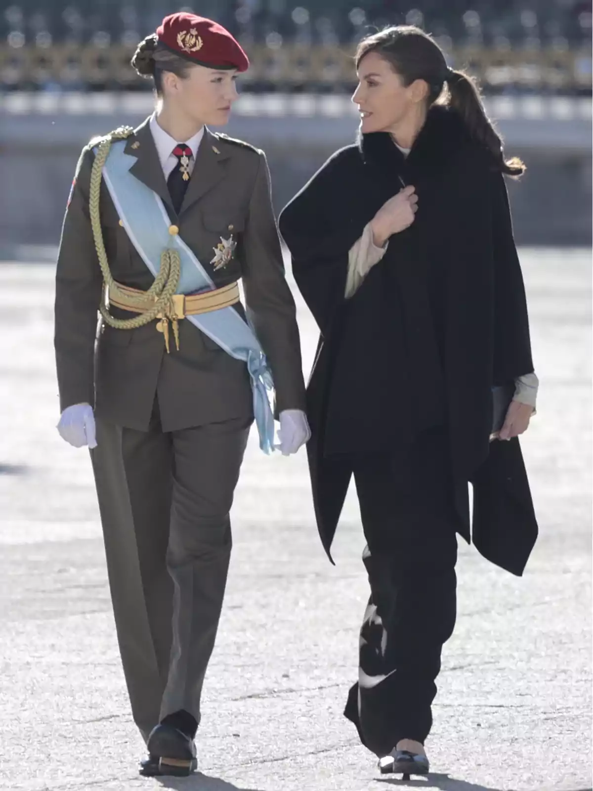 Dos mujeres caminando juntas, una de ellas vestida con uniforme militar y boina roja, mientras la otra lleva un abrigo negro.