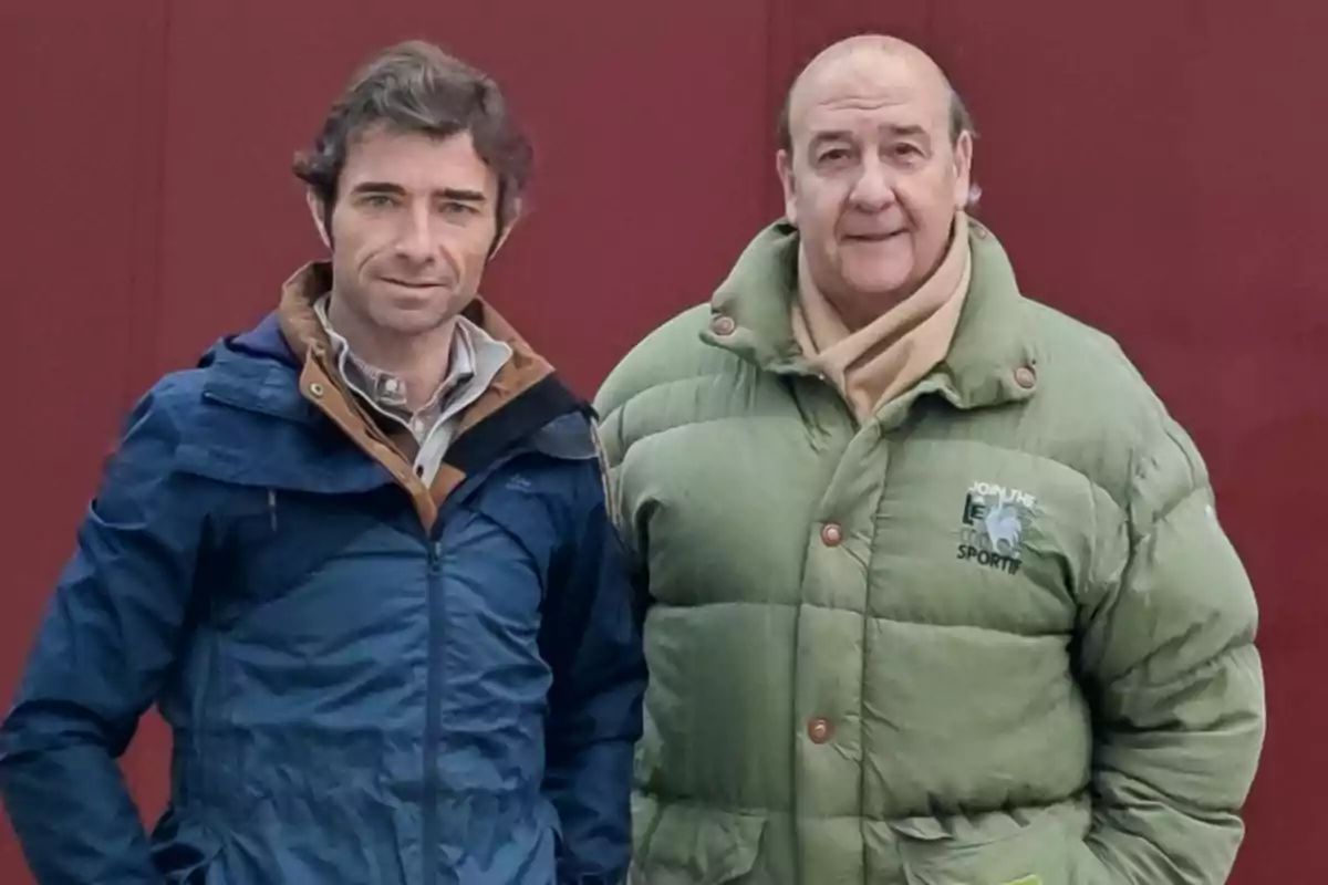 Dos hombres posando frente a una pared roja, uno con chaqueta azul y el otro con chaqueta verde.