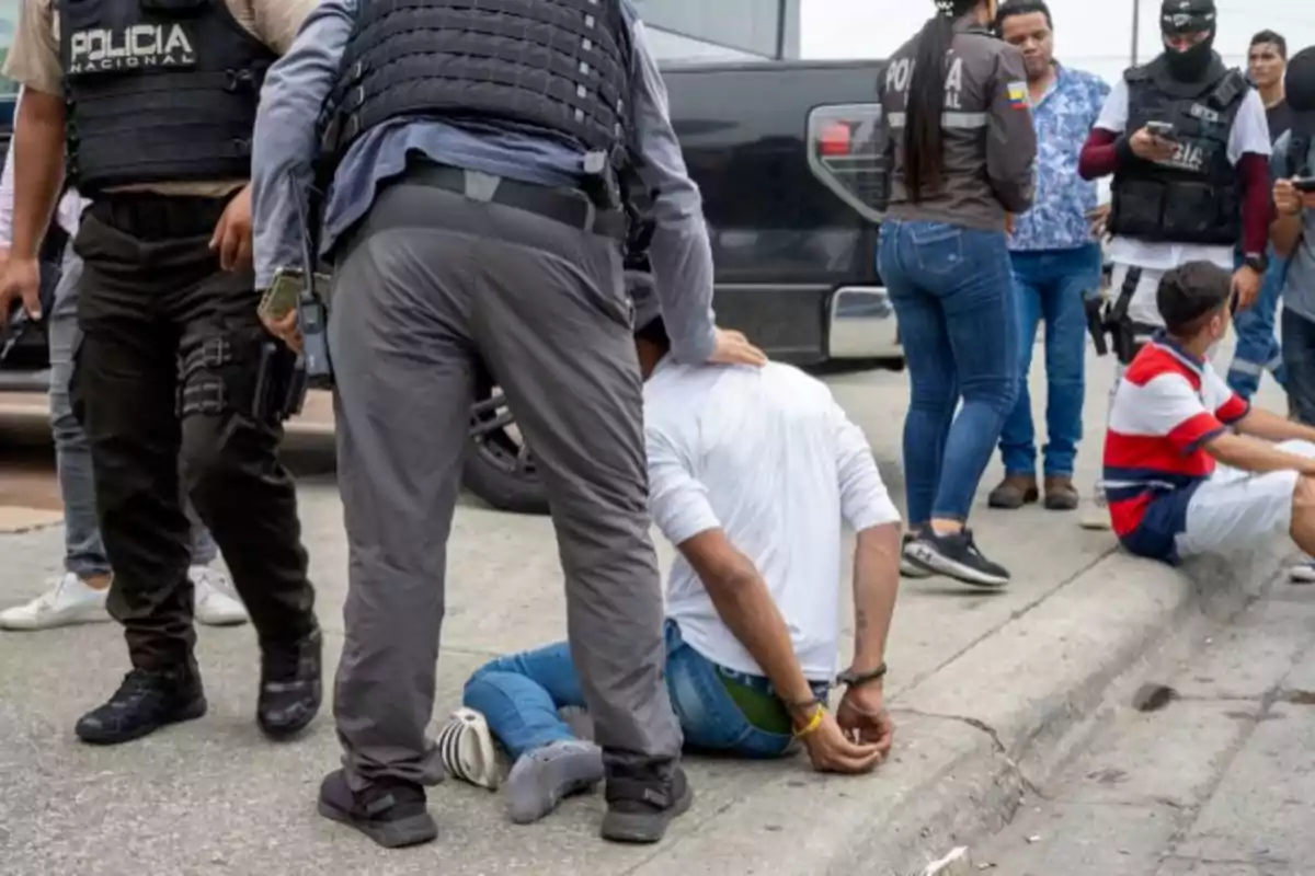 Policías detienen a varias personas en la calle, una de ellas está esposada y sentada en el suelo.