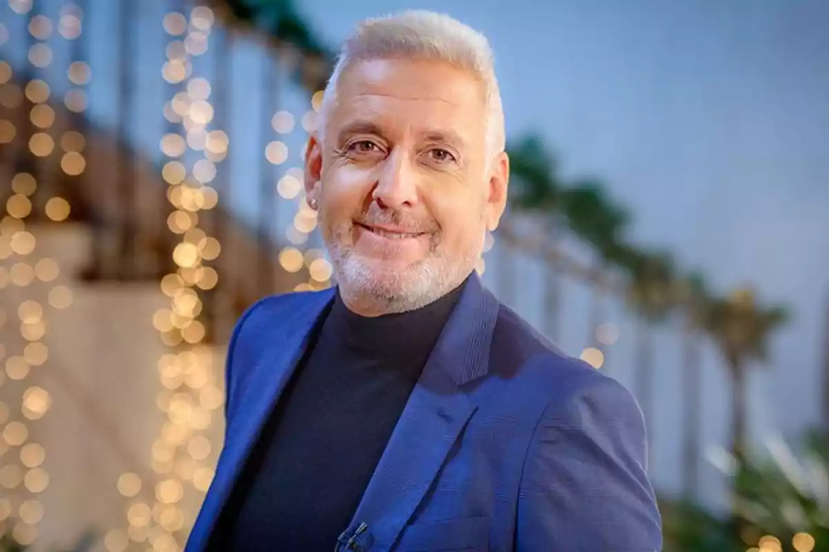 Hombre de cabello canoso y barba corta, vestido con un traje azul y suéter negro, sonriendo frente a un fondo con luces desenfocadas.