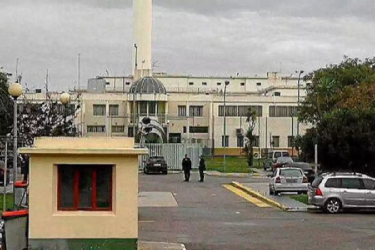 Edificio grande con una torre blanca en el centro, rodeado de un área de estacionamiento con varios autos y una caseta de vigilancia en primer plano.