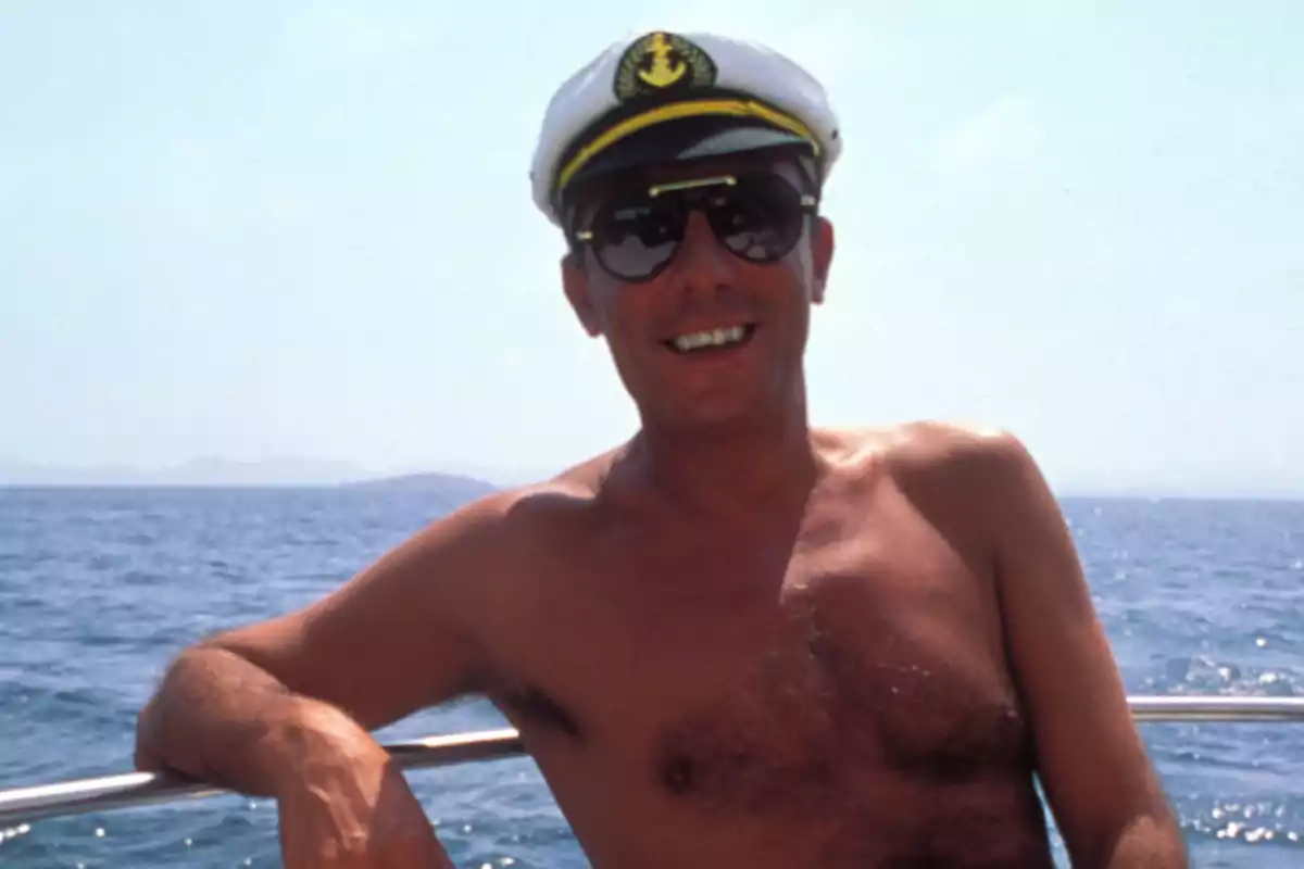 Hombre sin camisa con gafas de sol y gorra de capitán sonriendo en un barco con el mar de fondo.