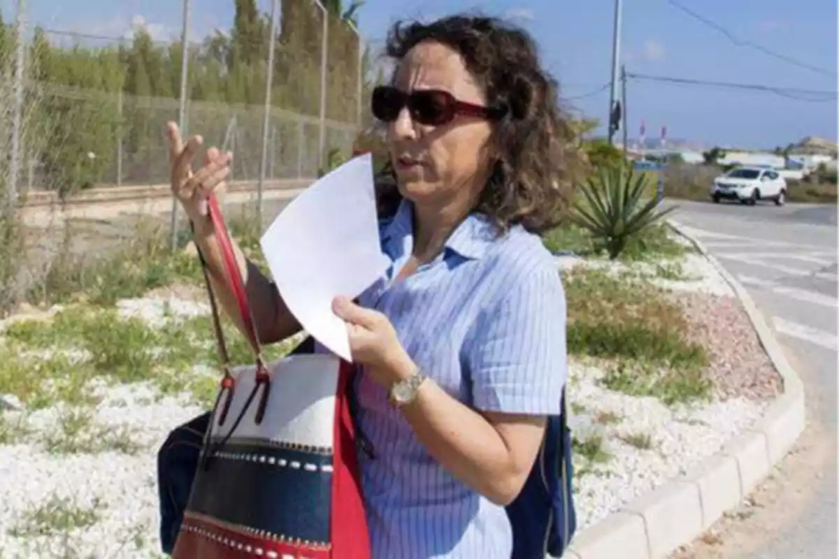 Una mujer con gafas de sol sostiene un papel mientras camina por una acera junto a una carretera, llevando un bolso de colores.