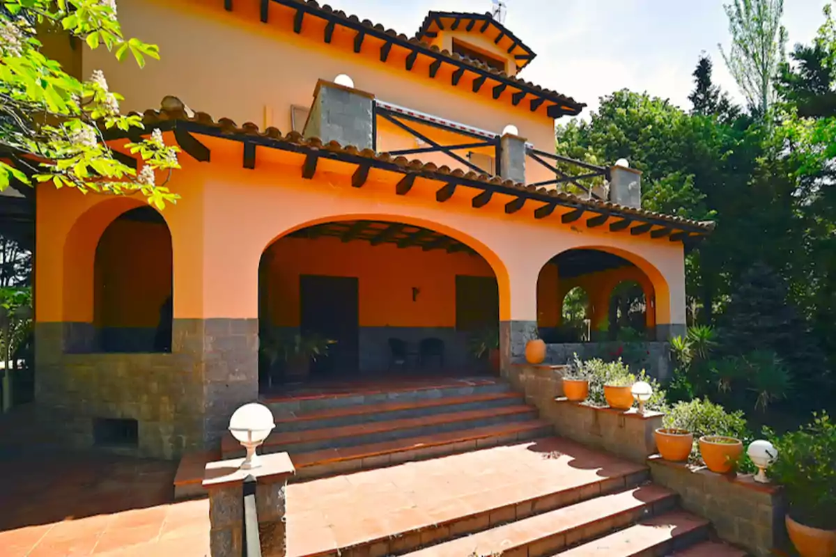 Casa de dos pisos con fachada de color naranja, techo de tejas y un porche con arcos y macetas.