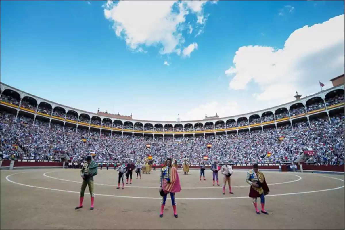 Plaza de toros llena de espectadores con toreros en el ruedo bajo un cielo azul con algunas nubes.